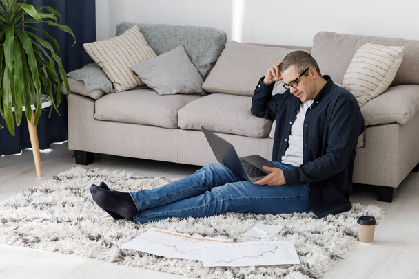 Un hombre vestido con ropa casual trabaja en casa, se sienta junto al sofá y trabaja con una computadora portátil. El hombre trabaja en un lugar remoto de trabajo - Foto, imagen