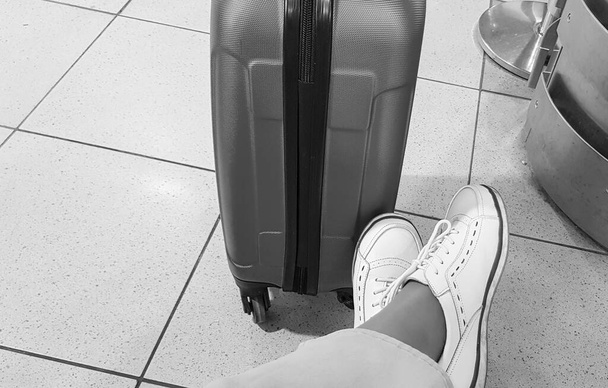 Pantolonlu ve spor ayakkabılı bir kadının bacağının siyah-beyaz bir fotoğrafı, rahat bir poz vermiş, bavulun yanına oturmuş, bir seyahat bekliyor.. - Fotoğraf, Görsel
