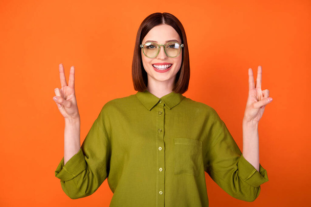 Photo of cool brunette hairdo millennial lady show v-sign wear specatcles green shirt isolated on orange color background - Zdjęcie, obraz