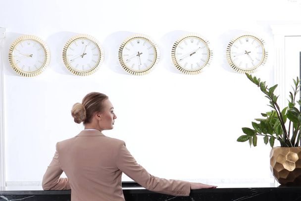 Girl in pink business suit stands at reception in hotel.Back view. Business travel concept. Vacation season. Clock is hanging on the wall.Travel or business trip.With copy space - Valokuva, kuva