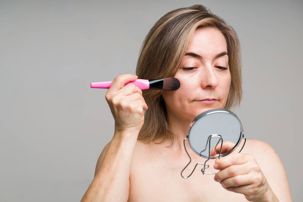 attrayant caucasien femme dans son 50 mettre blush et fondation avec une brosse de maquillage tout en regardant dans le miroir - Photo, image