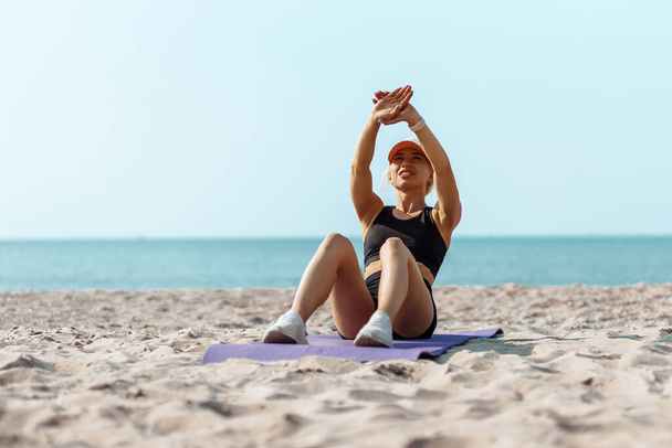 Sports young athlete on a yoga mat, with a slender body, working on the press, lying on the floor, training the press on the beach at sunrise - Photo, Image