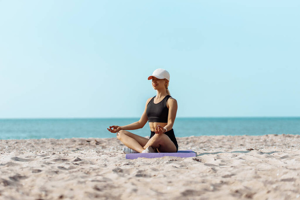 Mujer joven y deportiva haciendo yoga en la orilla del mar, mujer practicando yoga y meditación en posición de loto en la playa, hermosa mujer relajándose en la orilla del mar - Foto, imagen