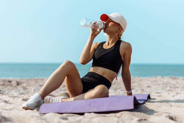 Fitness fiatal nő sportruházat ül és pihen edzés után, szabadban a strandon reggeli edzés után, pihenő és ivóvíz, fitness koncepció, egészséges, sport, életmód - Fotó, kép