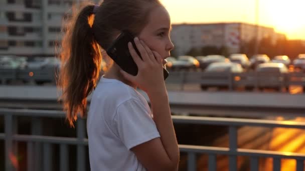Schoolgirl talks on smartphone with concentration at sunset - Footage, Video