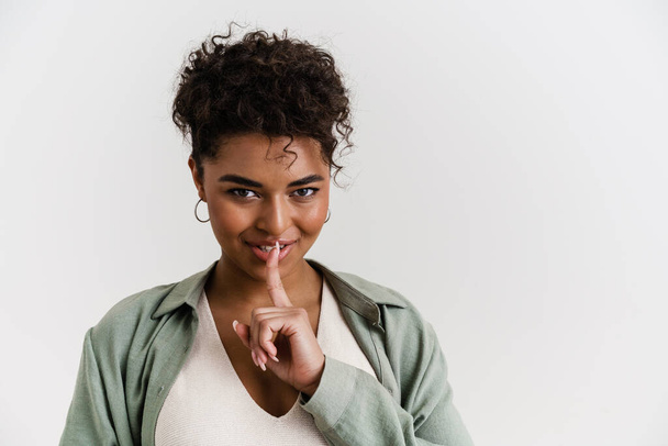 Young black woman in shirt smiling and showing silence gesture isolated over white background - Foto, immagini