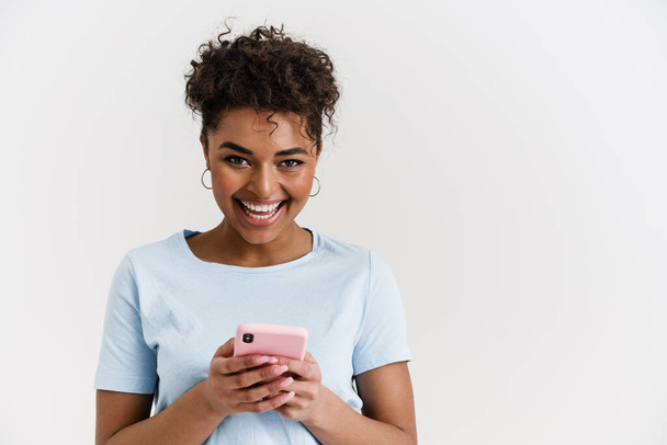 Smiling happy young african woman in casual wear standing over white wall, holding mobile phone - Foto, imagen
