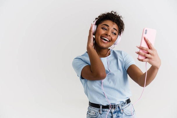 Laughing black woman listening music with headphones and cellphone isolated over white background - Foto, Imagen
