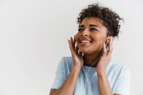 Happy casually dressed young african woman standing over white wall background, listening to music with earphones - Photo, image