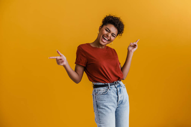 Happy casually dressed young african woman standing over yellow wall background, dancing - Foto, Imagem
