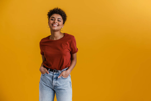 Happy casually dressed young african woman standing over yellow wall background, looking at camera - Foto, Imagem