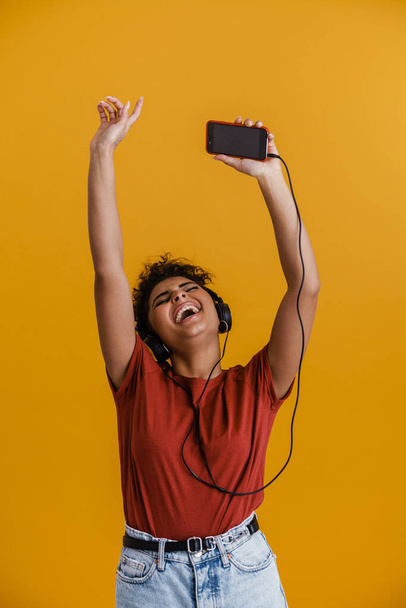 Laughing black woman listening music with headphones and cellphone isolated over yellow background - Φωτογραφία, εικόνα