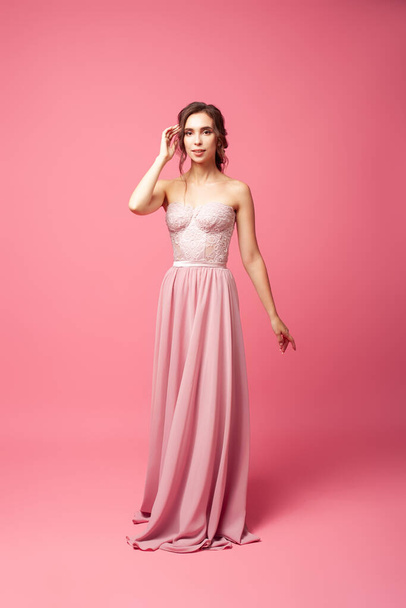 A young woman in a pink dress stands on an isolated background. A beautiful young woman in a long silk dress stands on a pink background - 写真・画像