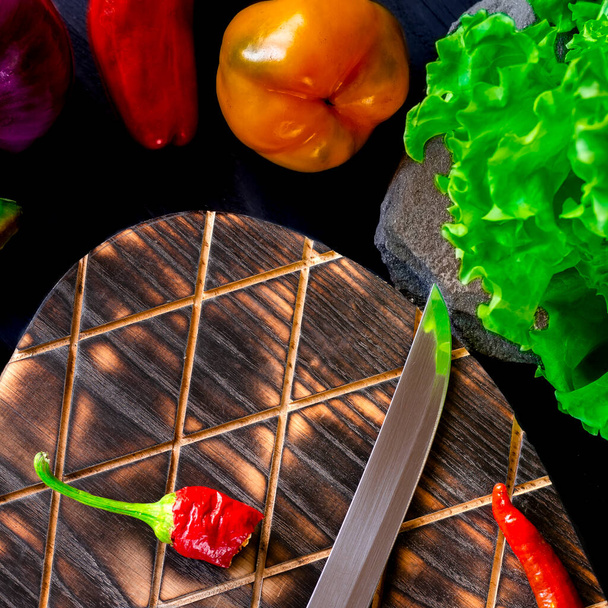 Poivrons rouges, jaunes, verts, chili, oignons bleus et laitue verte reposent sur une table en bois noir, à côté d'une planche à découper sur laquelle repose le poivre vert et un couteau avec un reflet de laitue verte - Photo, image