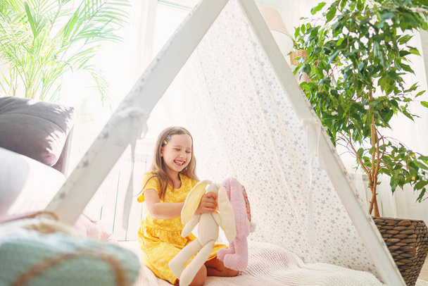 little cute girl is playing with the heroes of soft toys of rabbits sitting in a children's toy wigwam, the child is at home. Children's fantasy, happy childhood - Foto, imagen