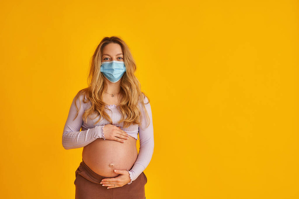 attractive pregnant woman in a mask, holding her stomach with her hand, stands straight and looks into the frame on a yellow background. Protection against viruses during pregnancy. Copy space - Photo, image
