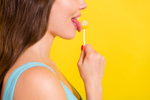 Cropped profile side view portrait of attractive girl licking lollipop isolated over vivid yellow color background - Zdjęcie, obraz