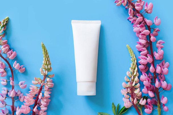 Unbranded white cosmetic squeeze tube and bright lupine flowers on blue background. Mockup, template. Container for body lotion, moisturizer cream, shower gel, shaving cream. - Foto, Imagen