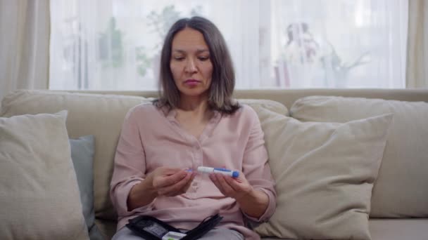 Mittlere Porträtaufnahme einer modernen, reifen Frau, die zu Hause auf dem Sofa sitzt und lernt, wie man Glukosemessgerät verwendet, telemedizinisches Konzept - Filmmaterial, Video