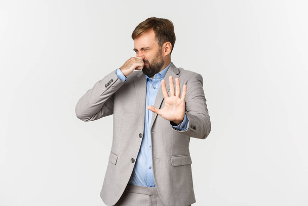 Portret zdegustowanego brodatego biznesmena w szarym garniturze, odrzucającego coś o okropnym zapachu, zamykającego nos i robiącego znak zaniku, stojącego nad białym tłem - Zdjęcie, obraz