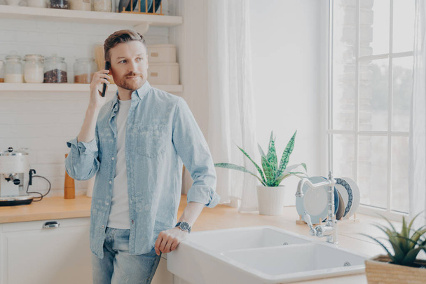 handsome young man in casual jeans shirt speaking on cellphone in kitchen - 写真・画像