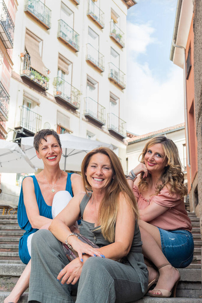Τρεις χαρούμενες ενήλικες γυναίκες κάθονται σε λιθοβολημένες σκάλες και κοιτάζοντας την κάμερα - Φωτογραφία, εικόνα