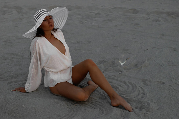 Full body verleidelijke vrouw in sexy shirt en hoed zitten sensueel op zandstrand in zonsondergang licht - Foto, afbeelding