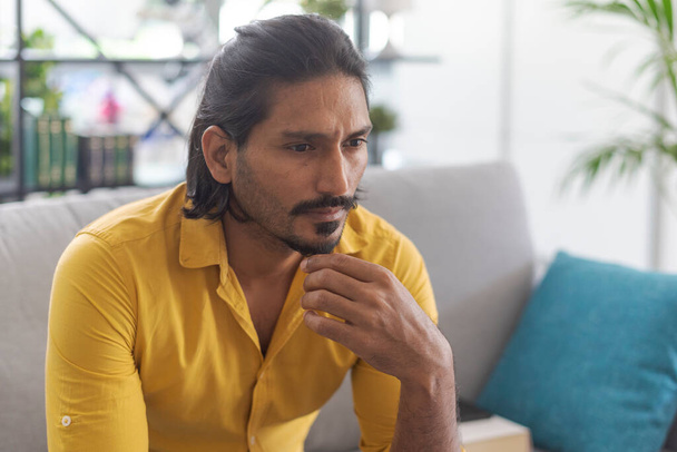 Indian man sitting on the couch at home and thinking with hand on chin - Photo, Image