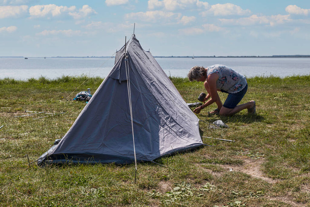 mature woman setting up a tent and putting down tent pegs on a field overlooking the water - Foto, afbeelding