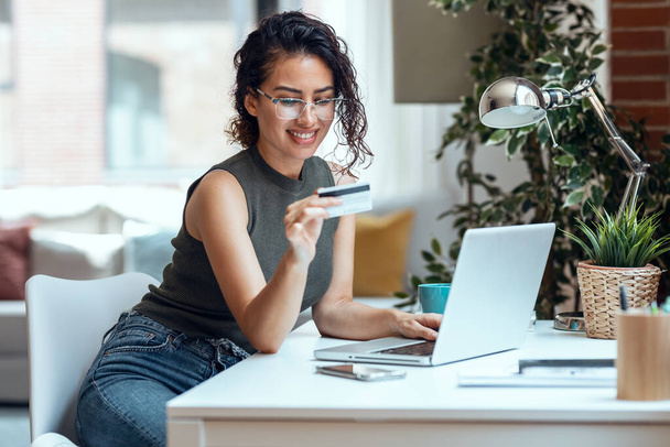Shot van schattige vrouw met witte creditcard voor het winkelen online met de computer, terwijl zitten in de woonkamer thuis. - Foto, afbeelding