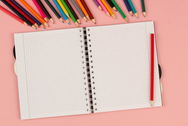 Open school squared notebook and color pencils on the pink background. Blank white sheet of paper book on the table. Office supplies on the desktop. Back to school. Copy space for text. Top view - Photo, Image