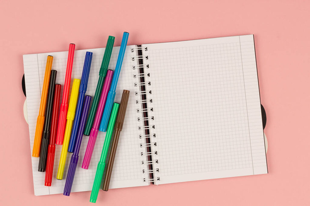 Open school squared notebook and colorful markers on the pink background. Blank white sheet of paper book on the table. Office supplies on the desktop. Back to school. Copy space for text. Top view - Photo, image