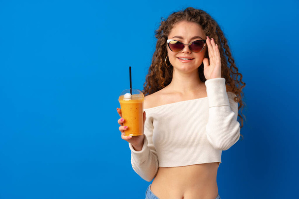 Beautiful curly-haired woman drinking orange juice against blue background - 写真・画像