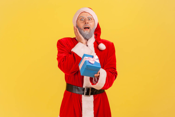 Extremely surprised elderly man with gray beard wearing santa claus costume holding wrapped blue present box, has gift on new year eve. Indoor studio shot isolated on yellow background. - Fotografie, Obrázek