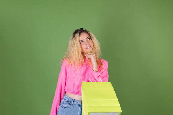 Mujer rubia joven con pelo rizado largo en suéter rosa sobre fondo verde con coloridas bolsas de papel feliz mirada alegre y reflexiva a un lado - Foto, imagen