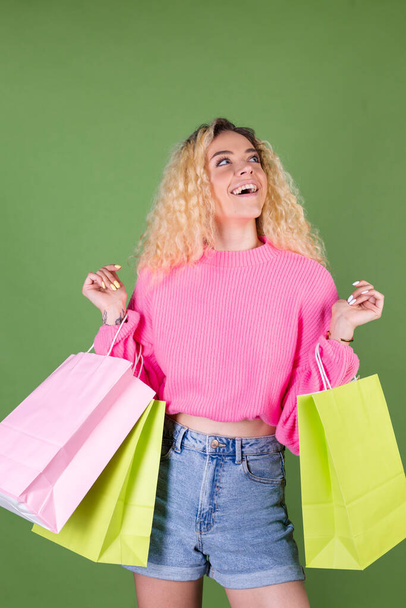 Young blonde woman with long curly hair in pink sweater on green background with colorful paper shopping bags happy cheerful excited - 写真・画像