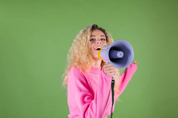 Mladá blondýna žena s dlouhými kudrnatými vlasy v růžovém svetru na zeleném pozadí šťastný pozitivní křik v megafonu požádat o pozornost  - Fotografie, Obrázek