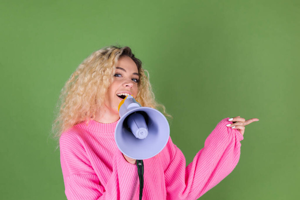 Jeune femme blonde avec de longs cheveux bouclés en pull rose sur fond vert heureux cris positifs en mégaphone doigt pointeur droit - Photo, image