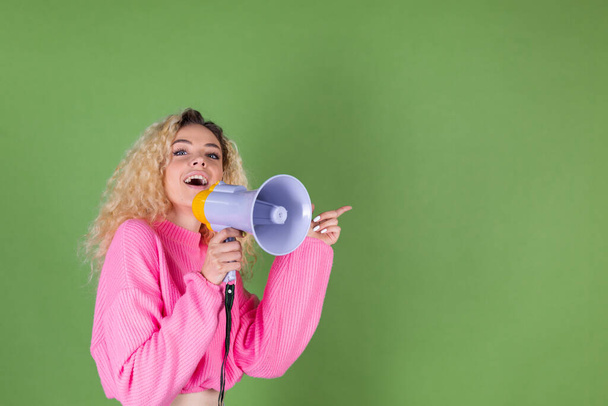 młoda blondynka kobieta z długimi kręconymi włosami w różowym swetrze na zielonym tle szczęśliwy pozytywne krzyki w megafon punkt palec prawo - Zdjęcie, obraz