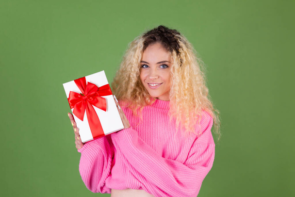 Young blonde woman with long curly hair in pink sweater on green background with gift box happy cheerful excited smiling - Foto, Imagen