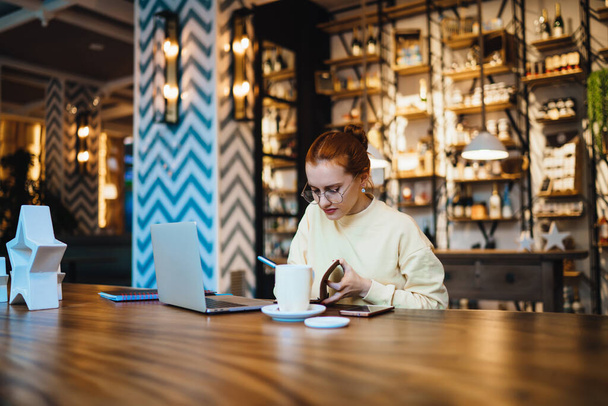 Pretty young woman in casual outfit and eyeglasses sitting at wooden table with mug of tea and mobile writing in notepad while browsing laptop in cafe in Barcelona city - Фото, изображение