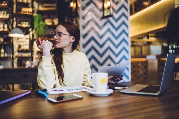 Brunette woman in yellow sweater and glasses with pink marker in hand looking away while sitting in cafe with laptop - Φωτογραφία, εικόνα