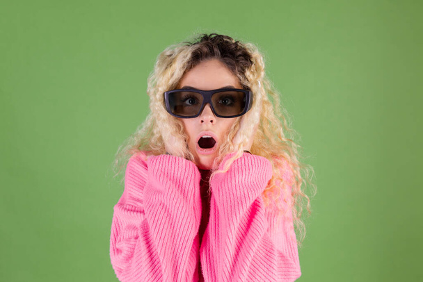 Young blonde woman with long curly hair in pink sweater on green background in 3d cinema shocked - Photo, Image