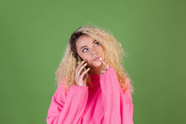 Young blonde woman with long curly hair in pink sweater on green background holding mobile phone thoughtful - Фото, зображення
