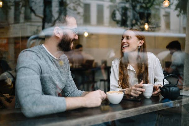 Happy young man and women in casual clothes smiling and talking at each other while sitting at table near window in modern cafe - Foto, Bild