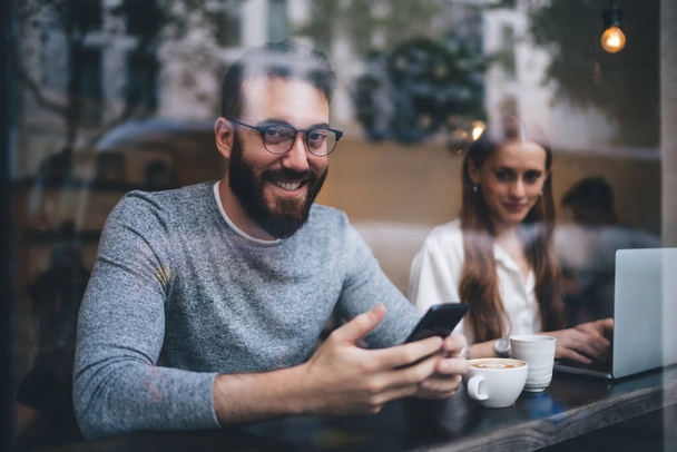 Young bearded man in casual wear and eyeglasses smiling at camera while browsing smartphone next to girlfriend working on project on laptop - Photo, image