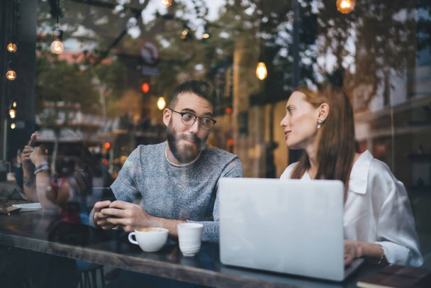 Young bearded man in eyeglasses typing on smartphone and ginger haired woman using laptop in casual clothes chilling at small table in cozy cafe with cups of hot beverage and talking to each other - Photo, Image