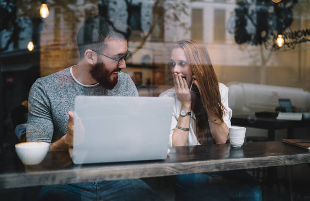 Surprised young couple in casual wear using laptop and enjoying coffee at bar counter in coffee shop sitting behind glass window overjoyed with new business opportunity - Fotoğraf, Görsel