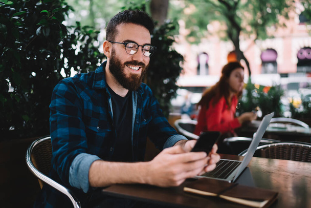 Happy male in eyeglasses and casual outfit smiling and browsing mobile phone while sitting at wooden table in street cafe - Fotoğraf, Görsel