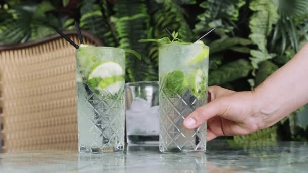 Hand legt zwei Cocktails Gin Tonic oder Mojito mit Gin, Minze, Eis und Limette auf den Tisch im Café. - Filmmaterial, Video